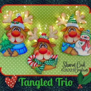 May include: Three painted wooden reindeer ornaments with festive hats and scarves. The reindeer are holding a Christmas tree, a candy cane, and a snowman. The ornaments are decorated with twinkling lights and the text "Tangled Trio" is visible at the bottom of the image.