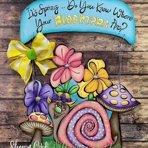 Op de afbeelding: Een houten bord met de tekst "It's Spring... Do You Know Where Your Bloomers Are?" Een slak met een roze en paarse schelp wordt omringd door bloemen en paddenstoelen. Het bord is versierd met een gele en witte strik.