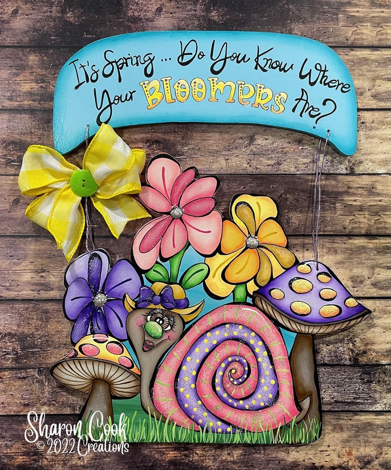 Op de afbeelding: Een houten bord met de tekst "It's Spring... Do You Know Where Your Bloomers Are?" Een slak met een roze en paarse schelp wordt omringd door bloemen en paddenstoelen. Het bord is versierd met een gele en witte strik.