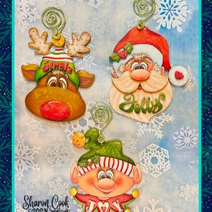 May include: Three Christmas decorations featuring a reindeer, Santa Claus, and an elf. The reindeer is brown with a red nose and a green hat with the word "Jingle" on it. Santa Claus is wearing a red hat and has a white beard. The elf is wearing a green hat and a red and white striped shirt. The decorations are all painted in a whimsical style.