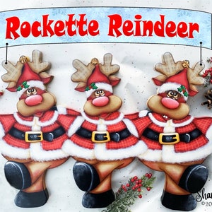 May include: Three wooden reindeer figurines dressed as Santa Claus, with red and white plaid coats, black belts, and white fur trim. The reindeer are standing in a row, with their heads tilted to the side and their mouths open. A banner above them reads "Rockette Reindeer".