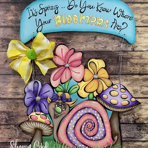Op de afbeelding: Een houten bord met de tekst "It's Spring... Do You Know Where Your Bloomers Are?" Een slak met een roze en paarse schelp, een strik en een groene neus is omringd door bloemen en paddenstoelen. Het bord is versierd met een gele en witte strik.