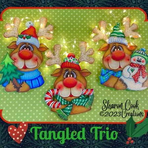May include: Three painted wooden reindeer ornaments with festive hats and scarves. The reindeer are holding a Christmas tree, a candy cane, and a snowman. The ornaments are decorated with twinkling lights and the text "Tangled Trio" is visible at the bottom of the image.