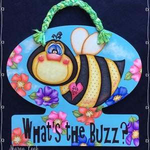 Op de afbeelding: Een blauwe en gele bij met een hartje waarop "Bee Kind" staat, is omringd door roze, paarse en witte bloemen. De bij bevindt zich op een blauwe ovaal met de tekst "What's the Buzz?" onderaan.