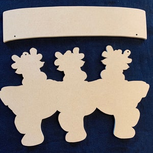 May include: Unpainted wooden craft kit featuring three reindeer silhouettes with a separate banner for a personalized message. The reindeer are standing side-by-side with their heads turned to the side. The banner is curved and has two holes for hanging.