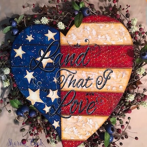 May include: A heart-shaped wooden decoration with a patriotic theme. The left side features a blue background with white stars, while the right side has red and white stripes. The words "Land That I Love" are written across the heart. The decoration is surrounded by a wreath of berries and greenery.
