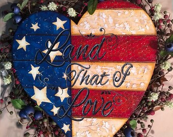 May include: A heart-shaped wooden decoration with a patriotic theme. The left side features a blue background with white stars, while the right side has red and white stripes. The words "Land That I Love" are written across the heart. The decoration is surrounded by a wreath of berries and greenery.