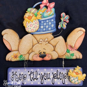 May include: A wooden sign with a purple background and white polka dots. The sign says "Hop 'til you plop!" with a yellow chick holding an egg on the right side. A brown bunny with a basket of Easter eggs on its head is above the sign.