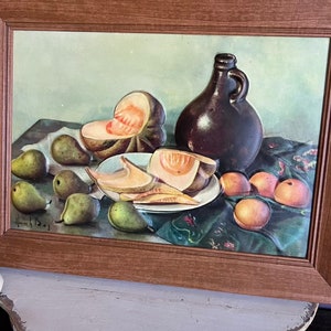 May include: A framed still life painting featuring a dark brown jug, sliced cantaloupe, pears, and peaches. The fruit is arranged on a green cloth. The painting is in a brown wooden frame.