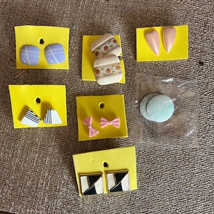 May include: A collection of earrings displayed on yellow cards. The earrings feature various designs, including geometric patterns, bows, and floral motifs. Colors include white, black, pink, and light blue. Some earrings are heart-shaped or square.