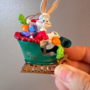 May include: A Bugs Bunny Christmas ornament. The ornament features Bugs Bunny in a green sleigh filled with carrots and gifts. The sleigh has gold accents and the word "BUGS" at the base. Bugs Bunny is wearing a Santa hat and red coat.