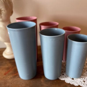 May include: Six vintage drinking glasses, four are light blue and two are pink. The glasses are arranged in a row on a wooden surface.