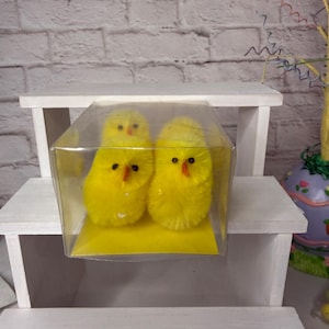 May include: Three fluffy yellow chicks with orange beaks and black eyes are displayed in a clear plastic box. The chicks are sitting on a yellow base. The box is on a white shelf, suggesting an Easter or spring theme.