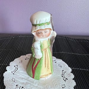 May include: A porcelain figurine of a shepherdess with a white bonnet, green and red robe, and a white lamb in her arms. She is holding a staff and has a closed-eye expression.