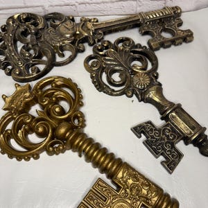 May include: Four ornate, antique-style keys in varying sizes and designs. The keys are primarily gold with bronze accents, featuring intricate scrollwork and decorative details. The keys are arranged on a white surface.