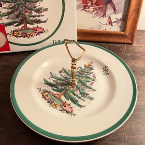 May include: A white ceramic serving plate with a green border and a gold handle. The plate features a Christmas tree design with colorful ornaments and wrapped gifts. A box with a matching design is in the background.