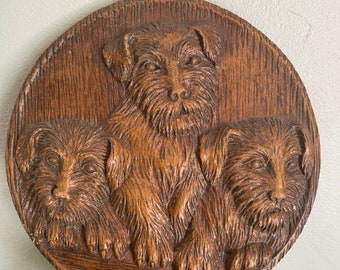 May include: A circular brown wall plaque featuring a detailed relief carving of three terrier dogs. The dogs are depicted with textured fur, pointed ears, and alert expressions. The plaque has a small hole at the top for hanging purposes.
