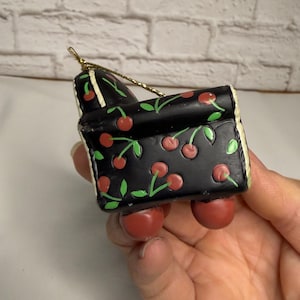 May include: A small, decorative ornament shaped like a miniature couch. The couch is black with a pattern of red cherries and green leaves. The ornament has red cherry-shaped feet and a gold string for hanging.