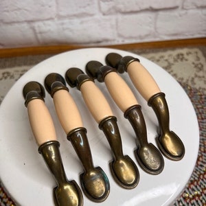 May include: Six antique-style cabinet pulls. Each pull features a cream-colored center, flanked by bronze-toned metal hardware. The pulls are arranged in a row on a white surface, suggesting a vintage or retro aesthetic.