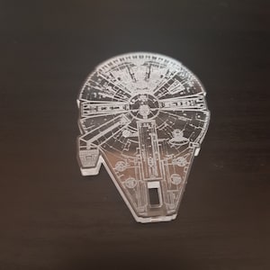 Könnte beinhalten: Ein durchsichtiger Acryl-Schlüsselanhänger mit einer detaillierten Umrisszeichnung des Raumschiffs Millennium Falcon aus Star Wars.
