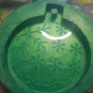Pode incluir: Um molde redondo verde com um design de floco de neve. O molde é feito de resina e é usado para criar ornamentos.