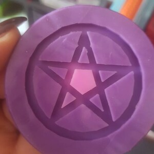 Pode incluir: Um objeto redondo roxo com um design de pentagrama em relevo. O objeto é de cor sólida com uma superfície lisa. O pentagrama, uma estrela de cinco pontas dentro de um círculo, é um símbolo frequentemente associado à Wicca e ao paganismo.