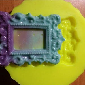 Puede incluir: Un molde de silicona para hacer manualidades de resina. El molde es amarillo y presenta un diseño de marco ornamentado detallado. El marco es morado y azul con un centro transparente.