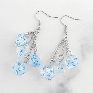 May include: Silver-tone earrings with clear dice featuring blue numbers. The dice are suspended from chains attached to the earring hooks. These earrings are designed for fans of tabletop role-playing games.