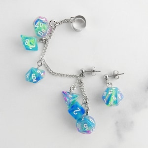 May include: Ear cuff and earring set featuring dice. The ear cuff has a silver-tone metal chain with several dice in shades of blue, green, and purple. The earring has a single die. The dice are numbered.