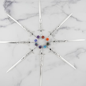 以下が含まれることがあります： 銀色の装飾的な剣が星型に配置されています。各剣には装飾的な柄と尖った刃があります。中央にはカラフルな宝石のようなビーズがあります。白い大理石の表面に配置されています。