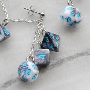 Op de afbeelding: Een paar zilveren oorhangers met een zilveren ketting en meerdere zwart-witte dobbelstenen met blauwe cijfers. De dobbelstenen zijn in een marmerpatroon.