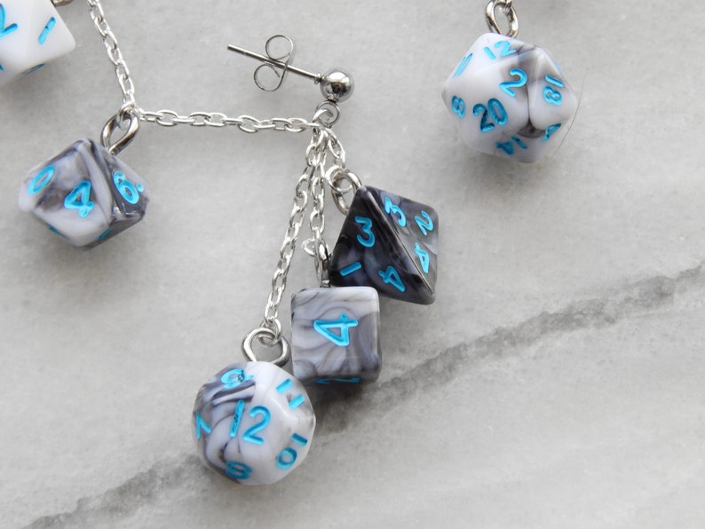 Op de afbeelding: Een paar zilveren oorhangers met een zilveren ketting en meerdere zwart-witte dobbelstenen met blauwe cijfers. De dobbelstenen zijn in een marmerpatroon.
