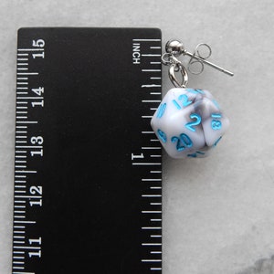 Op de afbeelding: Witte en grijze marmerachtige d20 dobbelsteen oorring met blauwe nummers. De oorring is ongeveer 2,5 cm lang en wordt getoond naast een liniaal.