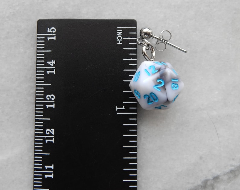 Op de afbeelding: Witte en grijze marmerachtige d20 dobbelsteen oorring met blauwe nummers. De oorring is ongeveer 2,5 cm lang en wordt getoond naast een liniaal.