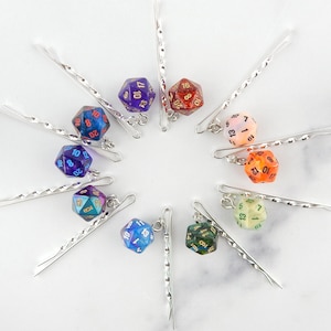 Op de afbeelding: Tien zilveren haarclips met iriserende dobbelstenen bedels. Elke dobbelsteen is van een andere kleur en heeft zwarte cijfers. De dobbelstenen zijn ongeveer 2,5 cm in diameter.