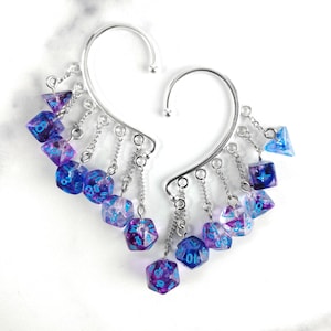 May include: A pair of silver-toned metal ear cuffs featuring multiple dangling miniature polyhedral dice. The dice are translucent with a purple and blue swirl pattern and bright blue numbers. Each cuff is connected to several small chains.