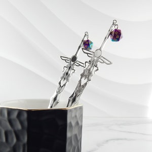 May include: Two silver-colored sword-shaped drink stirrers with decorative wings and diamond patterns. Each stirrer has a multi-colored 20-sided die attached. The stirrers are in a black and gold-rimmed mug.