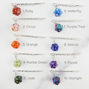 Op de afbeelding: Tien verschillende gekleurde d20 dobbelstenen bedels bevestigd aan zilveren haarclips. De dobbelstenen zijn gemaakt van helder hars met verschillende kleurwervelingen en glitter. De dobbelstenen zijn genummerd van 1 tot 20. De haarclips zijn zilverkleurig en hebben een golvend ontwerp.