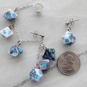 Op de afbeelding: Een paar oorringen met een zilveren ketting met meerdere dobbelsteenbedels. De dobbelstenen zijn wit met zwarte marmering en blauwe cijfers. De oorringen worden getoond naast een Nederlandse euro voor referentie van de grootte.