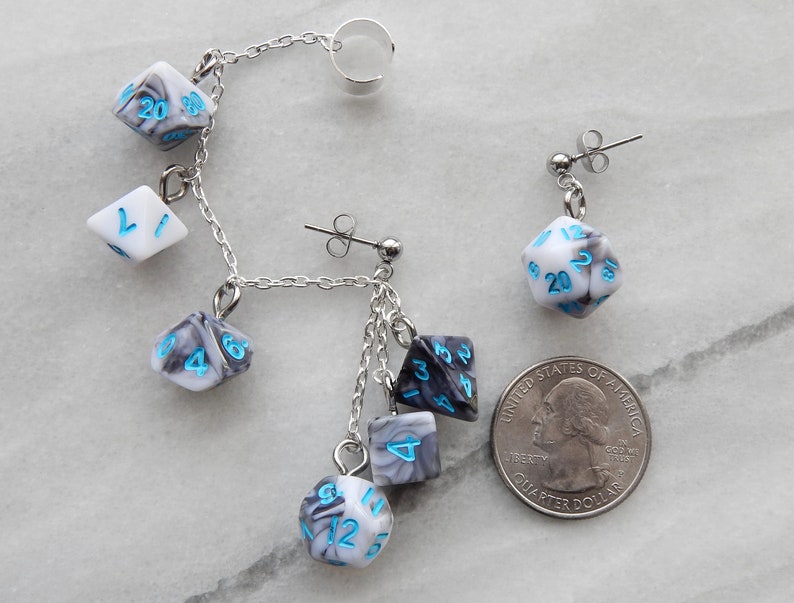 Op de afbeelding: Een paar oorringen met een zilveren ketting met meerdere dobbelsteenbedels. De dobbelstenen zijn wit met zwarte marmering en blauwe cijfers. De oorringen worden getoond naast een Nederlandse euro voor referentie van de grootte.