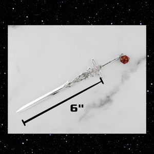 May include: A silver sword hair stick with a red and gold dice charm. The hair stick is 6 inches long.