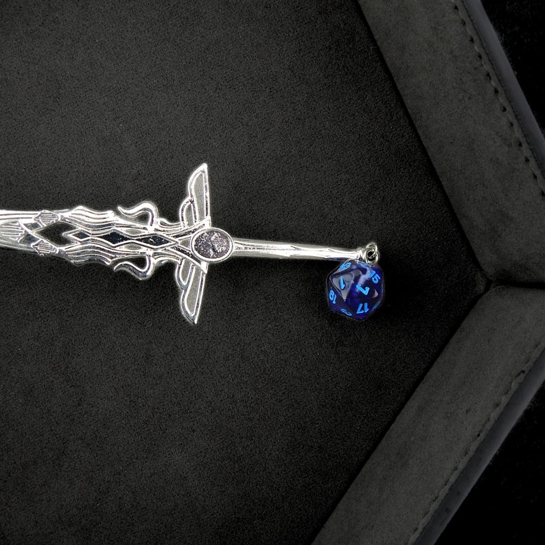 May include: A silver-colored sword-shaped pendant with intricate detailing and a blue, multi-sided die charm. The pendant has a decorative hilt and blade design. The die is dark blue with white numbers.