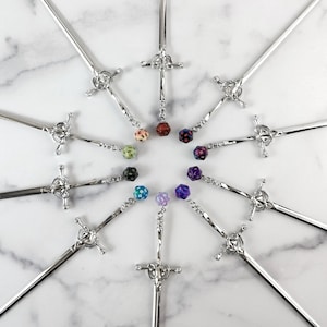 May include: A collection of silver letter openers, each designed as a sword. Each opener has a decorative handle and is adorned with a colorful, patterned dice charm. The openers are arranged in a circular pattern on a marble surface.