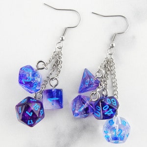 May include: Dangle earrings with multiple purple and blue polyhedral dice. The dice are connected by silver chains and have blue numbering. The earrings have silver-coloured fish hook ear wires.