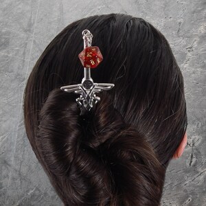 May include: A silver hair pin with a sword design, topped with a red and gold D20 die. The hair accessory is inserted into a bun of dark hair. The pin has an ornate design and is set against a gray background.