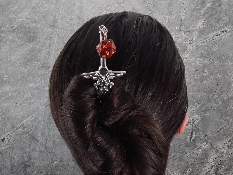 May include: A silver hair pin with a sword design, topped with a red and gold D20 die. The hair accessory is inserted into a bun of dark hair. The pin has an ornate design and is set against a gray background.