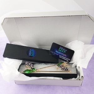 May include: An open white box reveals a gold-colored hair stick with green enamel leaves and purple dice accents. A black box with the text "LEVELED UP GEEK" and a business card are also visible. The business card has the text "LEVELED UP GEEK" and the designer's name.
