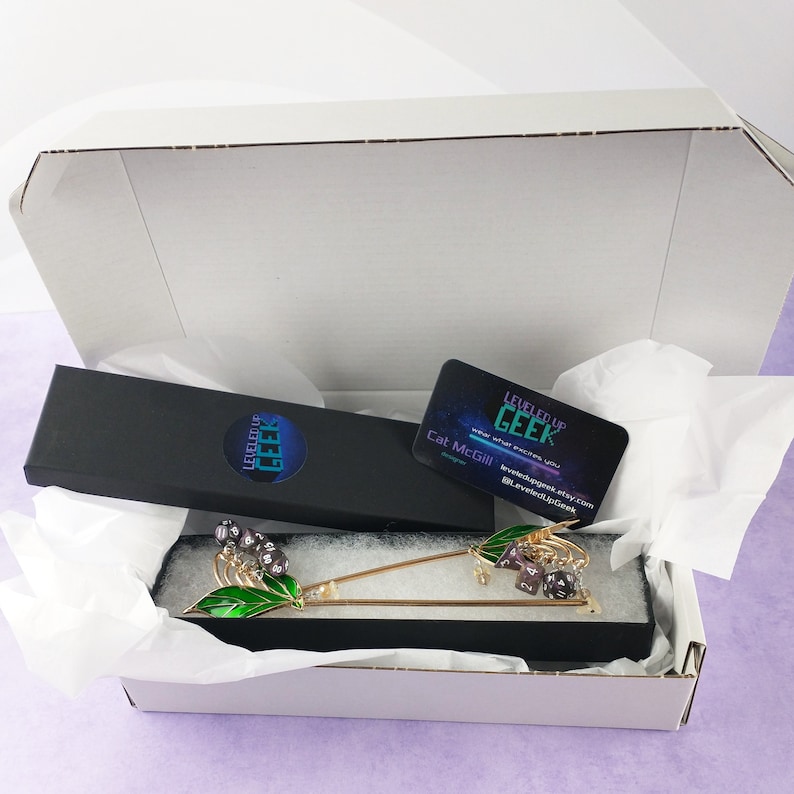 May include: An open white box reveals a gold-colored hair stick with green enamel leaves and purple dice accents. A black box with the text "LEVELED UP GEEK" and a business card are also visible. The business card has the text "LEVELED UP GEEK" and the designer's name.