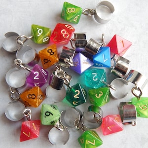 May include: A selection of colourful polyhedral dice charms in shades like green, red, and purple. Each die is connected to a silver-tone metal ring. The dice display numbers in gold. These charms are suitable for jewellery or crafting projects.
