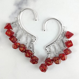 May include: A pair of silver-toned ear cuffs featuring dangling red glitter polyhedral dice with gold-coloured numbers. Each cuff is adorned with multiple miniature dice of various shapes, including d4, d6, d8, d10, d12, and d20, suspended by small silver-toned metal chains.